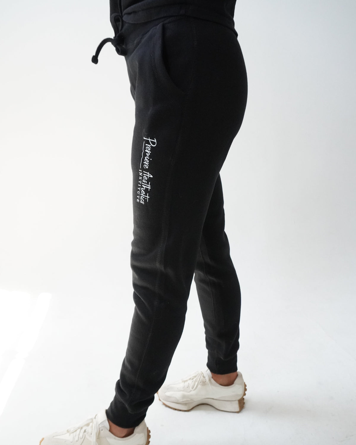 Black jogger sweatpants with white vertical script text on the left leg, shown on a model from the waist down wearing white sneakers against a white studio background.