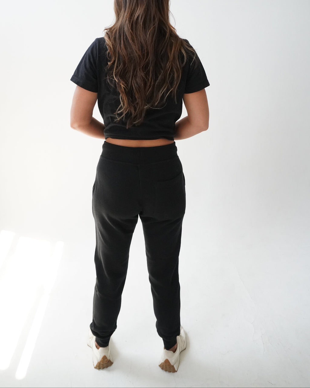 A woman viewed from behind wearing a black short-sleeve crop top and black jogger pants, standing against a plain white studio background.