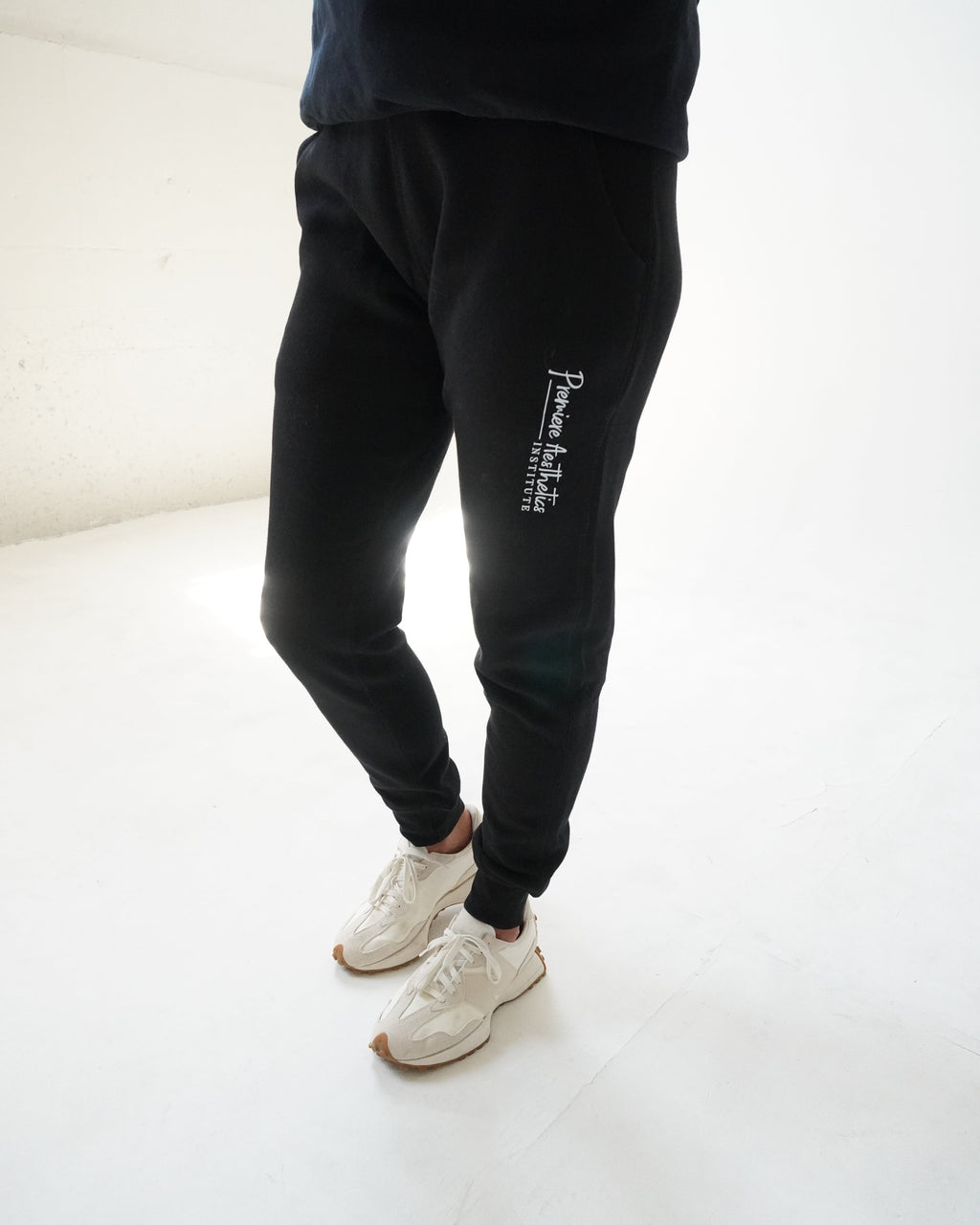 A person wearing black jogger sweatpants with white vertical text on the left leg, paired with white sneakers, standing in a bright white studio space.