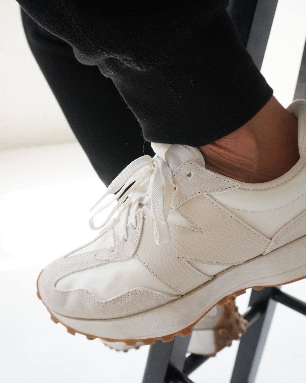 A close-up of a person's foot wearing a cream-colored leather sneaker with a brown gum sole, sitting on a black metal stool.